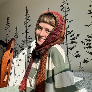 May include: A person wearing a striped green and white shirt with a red, orange, and green crocheted scarf and hat. The scarf and hat have tassels.