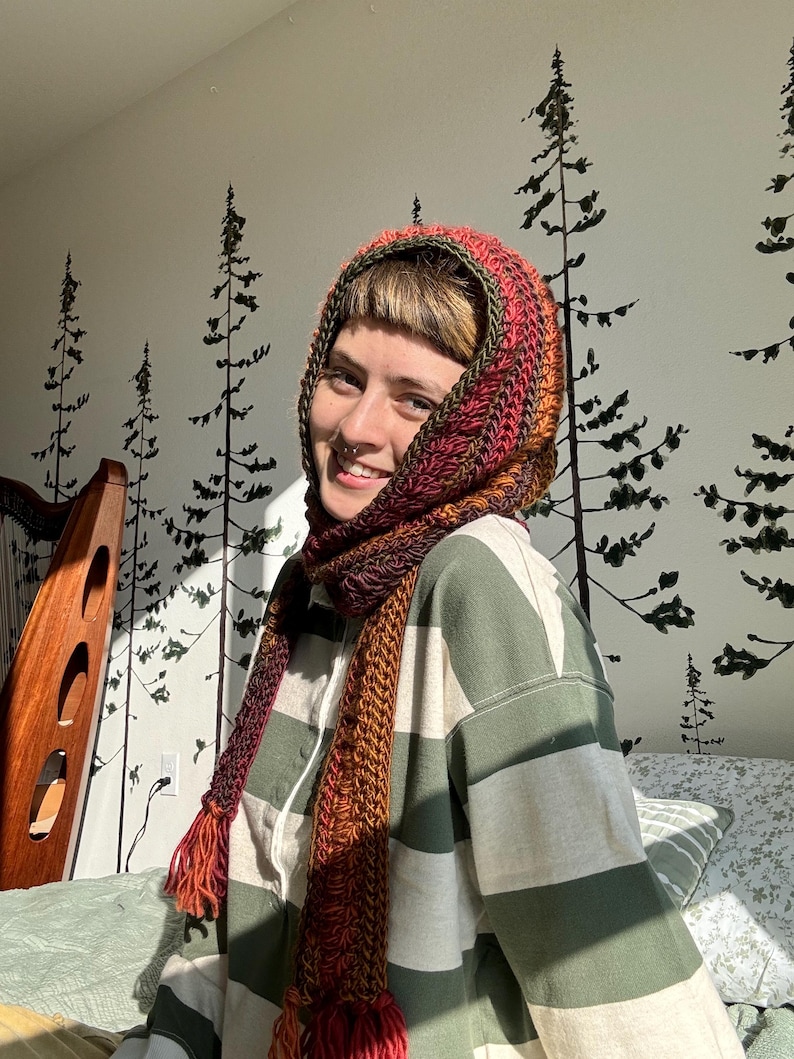 May include: A person wearing a striped green and white shirt with a red, orange, and green crocheted scarf and hat. The scarf and hat have tassels.