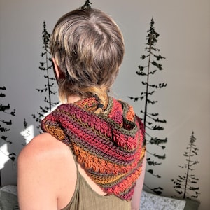 May include: A person wearing a crocheted hooded scarf in shades of orange, red, and green. The scarf is worn over a green tank top.