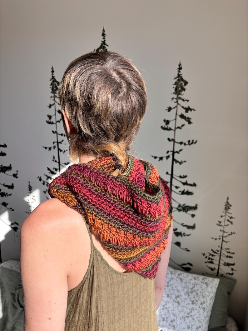 May include: A person wearing a crocheted hooded scarf in shades of orange, red, and green. The scarf is worn over a green tank top.