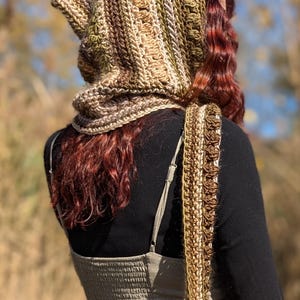 May include: A crocheted hooded scarf in shades of brown, beige, and green. The scarf is worn by a person with long, curly red hair. The scarf is long enough to be worn as a shawl.