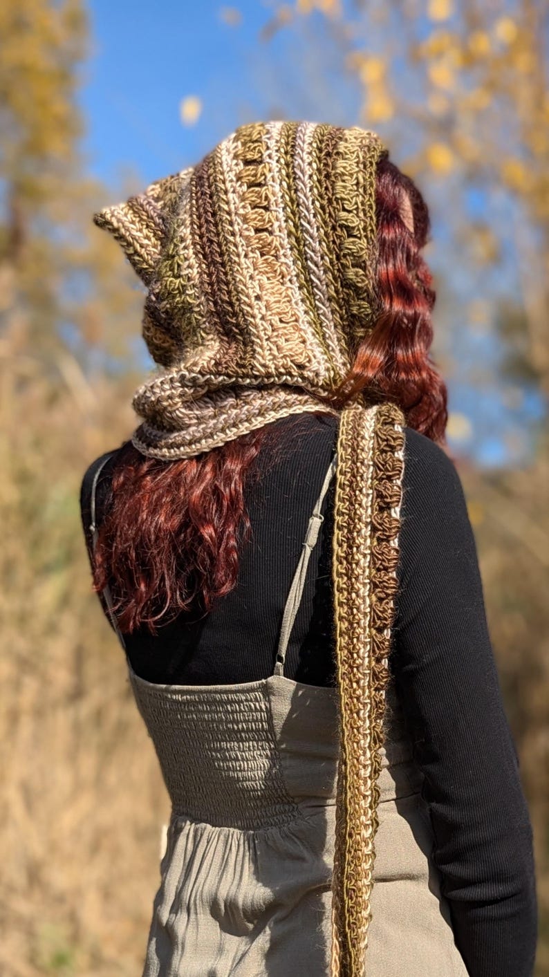 May include: A crocheted hooded scarf in shades of brown, beige, and green. The scarf is worn by a person with long, curly red hair. The scarf is long enough to be worn as a shawl.