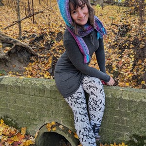May include: A woman wearing a hooded scarf with a gradient of purple, blue, and turquoise. The scarf has a tassel on each end. She is wearing a gray dress and black and white patterned leggings. She is sitting on a brick wall in a forest setting with fallen leaves.