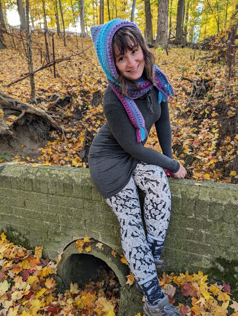 May include: A woman wearing a hooded scarf with a gradient of purple, blue, and turquoise. The scarf has a tassel on each end. She is wearing a gray dress and black and white patterned leggings. She is sitting on a brick wall in a forest setting with fallen leaves.