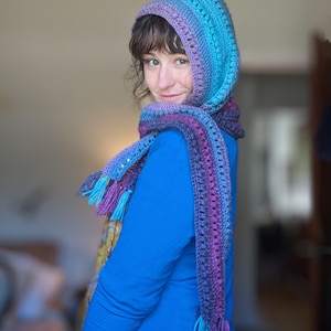 May include: A hooded scarf with a long fringe in shades of purple, blue, and teal. The scarf is crocheted and has a textured pattern.