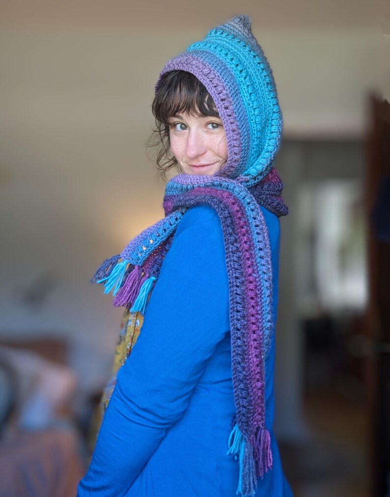 May include: A hooded scarf with a long fringe in shades of purple, blue, and teal. The scarf is crocheted and has a textured pattern.
