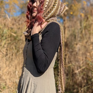 May include: A woman wearing a crocheted hooded scarf in shades of brown, green, and cream. The scarf has a long fringe and is worn with a long, beige dress and a black long-sleeved shirt.