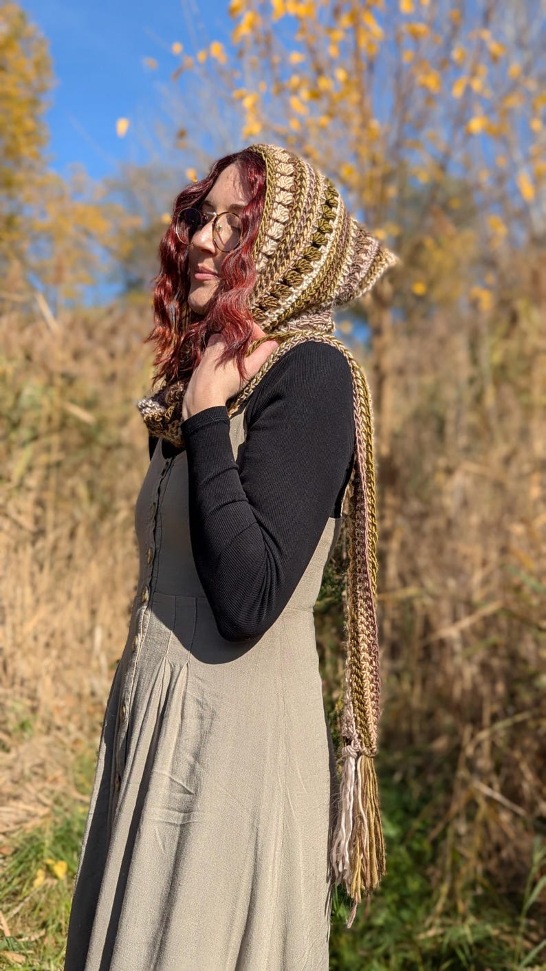 May include: A woman wearing a crocheted hooded scarf in shades of brown, green, and cream. The scarf has a long fringe and is worn with a long, beige dress and a black long-sleeved shirt.