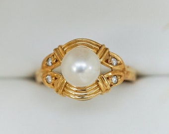 May include: A gold ring with a large, round pearl center stone. The ring has a decorative, woven-style design around the pearl and small accent stones. The ring is displayed in a white jewelry box.