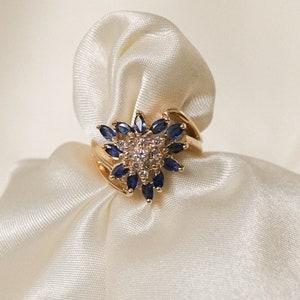 May include: A gold ring with a large blue gemstone surrounded by smaller clear gemstones. The ring is set on a white satin fabric.