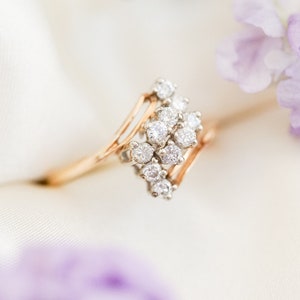 Pode incluir: Um delicado anel de ouro com um grupo de diamantes brilhantes engastados em uma banda delicada de v&aacute;rias camadas. O anel &eacute; fotografado em um fundo branco com flores roxas.