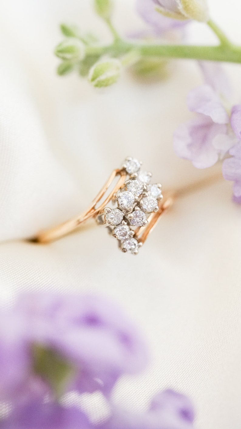 Pode incluir: Um delicado anel de ouro com um grupo de diamantes brilhantes engastados em uma banda delicada de v&aacute;rias camadas. O anel &eacute; fotografado em um fundo branco com flores roxas.