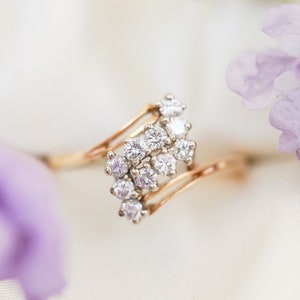 Pode incluir: Um anel de ouro com um aglomerado de pequenos diamantes engastados em um design em espiral. O anel &eacute; fotografado em um fundo branco com flores roxas.