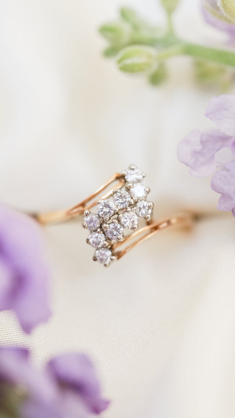 Pode incluir: Um anel de ouro com um aglomerado de pequenos diamantes engastados em um design em espiral. O anel &eacute; fotografado em um fundo branco com flores roxas.