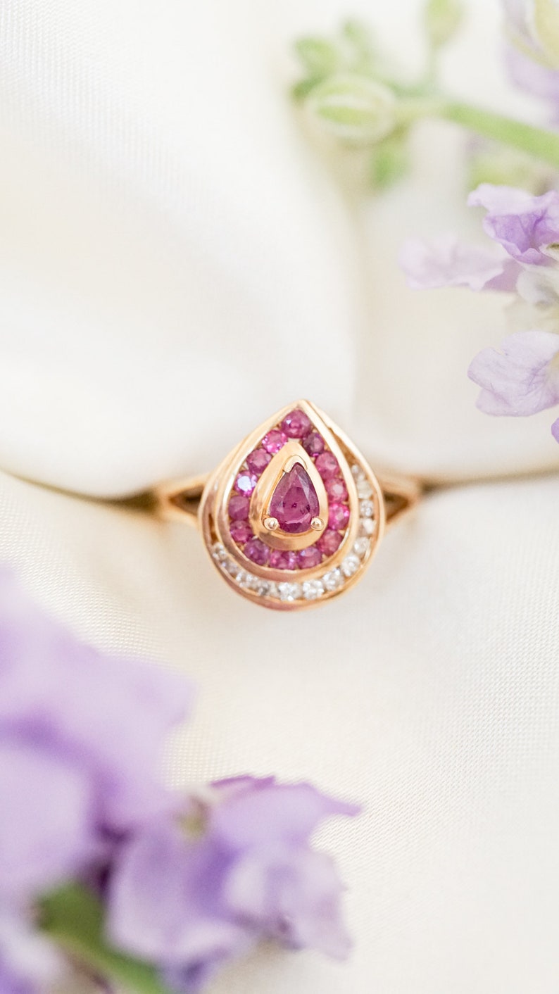 May include: A pear-shaped ring with a pink gemstone surrounded by smaller pink gemstones and a halo of white gemstones. The ring is set in gold.