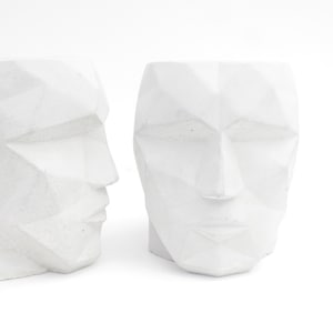 May include: Two white geometric face planter pots. The planters are made of concrete and have a textured surface. The planters are in the shape of a human face with a geometric design.