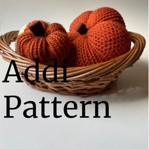 Pode incluir: Duas abóboras de malha laranja em uma cesta de vime. O texto "Addi Pattern" é visível abaixo da cesta.