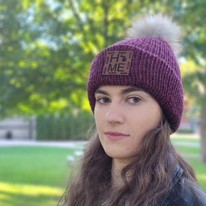 May include: A deep purple knit beanie with a faux fur pom-pom. The hat features a brown leather patch with the word "HOME" and a state outline. The hat is worn by a person.