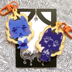 May include: Two blue cat-shaped keyrings with orange clasps. The cats have sad expressions and are surrounded by a yellow flame design. The background is a black and white abstract pattern.