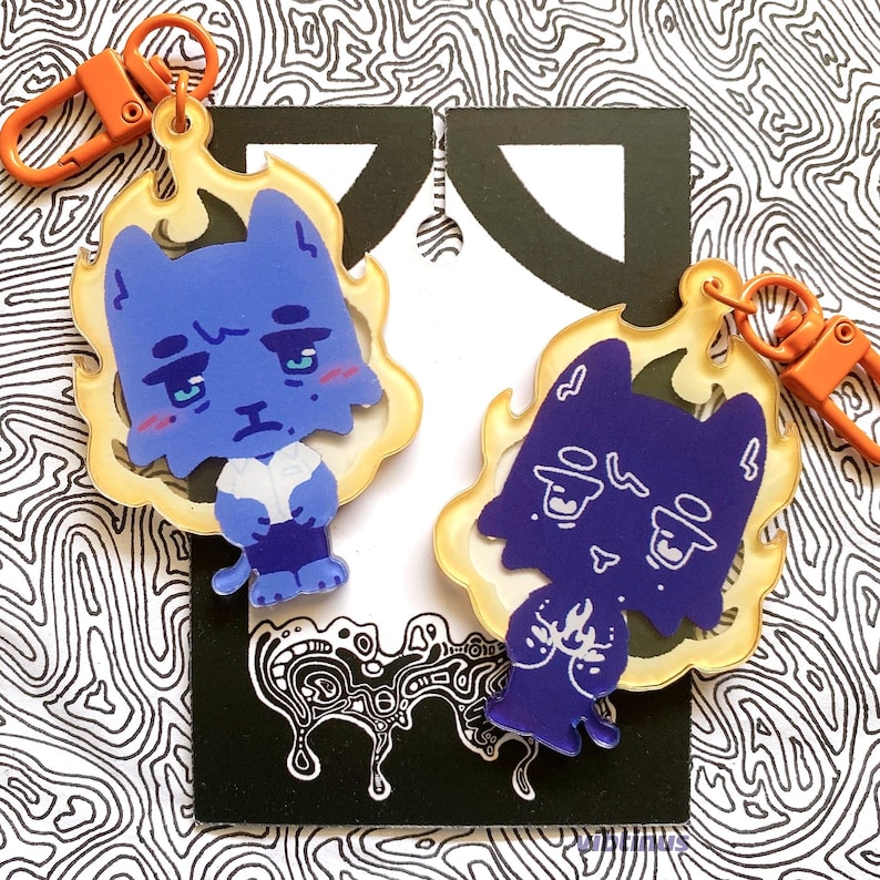 May include: Two blue cat-shaped keyrings with orange clasps. The cats have sad expressions and are surrounded by a yellow flame design. The background is a black and white abstract pattern.
