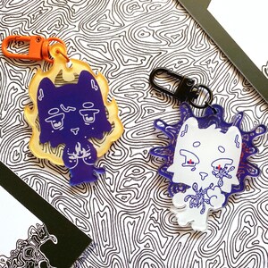 May include: Two acrylic keychains featuring cartoon cat designs. One is purple with a yellow flame border and an orange clip. The other is white with a blue splatter design and a black clip. The background has a black and white pattern.