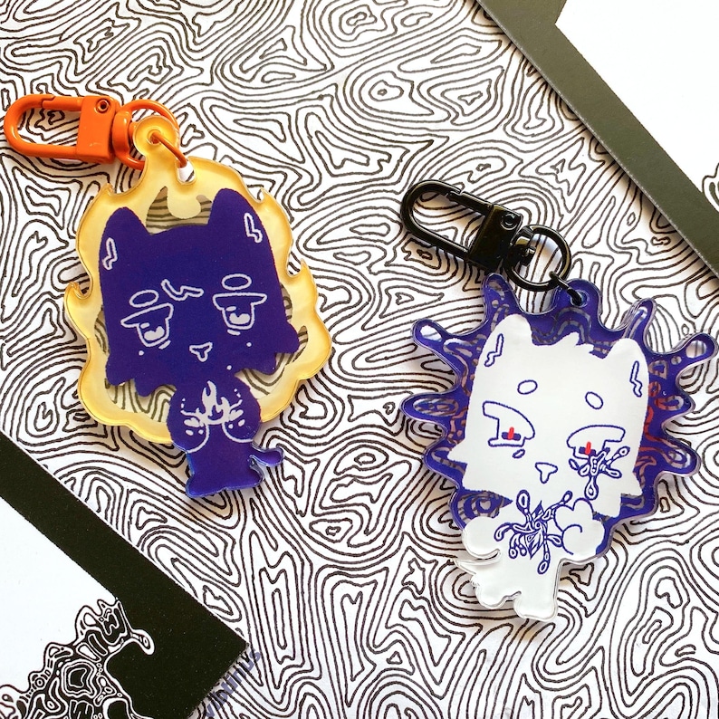 May include: Two acrylic keychains featuring cartoon cat designs. One is purple with a yellow flame border and an orange clip. The other is white with a blue splatter design and a black clip. The background has a black and white pattern.