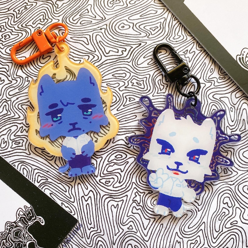 May include: Two cartoon cat keyrings. One is blue with a yellow flame-like border and an orange clasp. The other is white with a purple spiky border and a black clasp. Both have a sad expression and are on a black and white patterned surface.