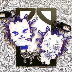 May include: Two acrylic keychains with a white cat design, blue and red details, and a purple splatter effect. Each keychain has a black clasp. The background features a black and white abstract pattern. The keychains are ideal for bags or keys.