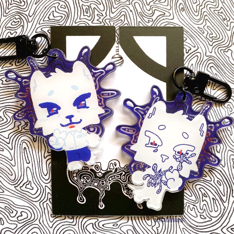 May include: Two acrylic keychains with a white cat design, blue and red details, and a purple splatter effect. Each keychain has a black clasp. The background features a black and white abstract pattern. The keychains are ideal for bags or keys.