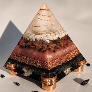 May include: A layered pyramid sculpture with a clear top, white stones, and copper wire accents. The pyramid has a dark red layer, a copper mesh layer, and a black base with copper-colored feet. Small black stones surround the base.