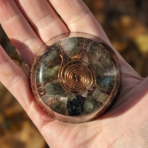 Op de afbeelding: Een ronde orgone-energiegenerator van heldere hars met een koperen spiraal en ingebedde groene en grijze stenen. Het apparaat wordt in een open hand gehouden. De achtergrond is een vage herfstbladeren.