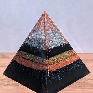 Op de afbeelding: Een gelaagde piramidesculptuur met een zwarte basis, met banden van goud-, koper- en zilverkleurige materialen. De top is een koperkleurig draadframe. De piramide wordt tentoongesteld op een houten oppervlak.