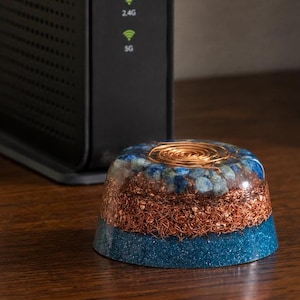 Pode incluir: Uma pirâmide de orgone multicamadas com uma bobina de cobre no topo. A pirâmide apresenta uma base azul, uma camada intermediária marrom e uma camada superior com pedras azuis. A pirâmide está em uma superfície de madeira.