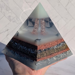 Könnte beinhalten: Eine mehrschichtige Orgonpyramide. Die Pyramide hat eine hellblaue Harzaußenseite mit Kupferspiralen und Schichten aus metallischen Materialien in Kupfer-, Silber- und Goldtönen. Die Pyramide wird in einer Hand gehalten.