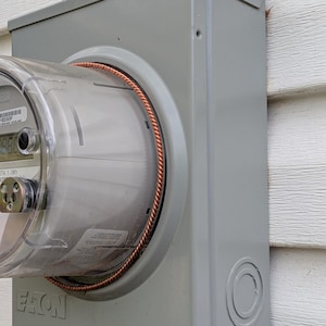 May include: A gray metal electrical meter box with a clear plastic meter inside. The meter has a copper wire wrapped around it. The box has a circular opening on the right side.