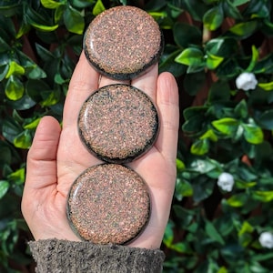 Peut inclure: Trois pierres rondes et polies avec une surface texturée de couleur cuivrée sont tenues dans une main. Les pierres ont une bordure sombre et une finition brillante. L'arrière-plan est un vert luxuriant avec des fleurs blanches.