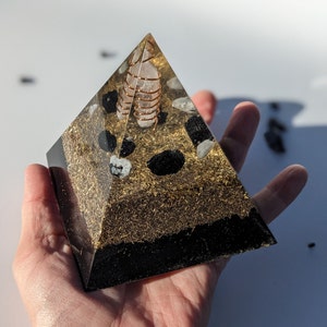 May include: A clear pyramid with a gold and black speckled base. Inside the pyramid is a clear quartz crystal with a copper wire wrapped around it. The pyramid is sitting on a hand.