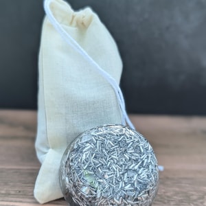 May include: A clear, spherical object filled with silver-coloured, shard-like pieces. It sits in front of a small, beige drawstring bag. The background is a dark grey, and the surface is a light brown wood.