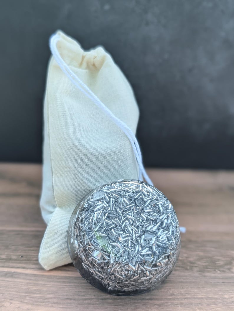 May include: A clear, spherical object filled with silver-coloured, shard-like pieces. It sits in front of a small, beige drawstring bag. The background is a dark grey, and the surface is a light brown wood.