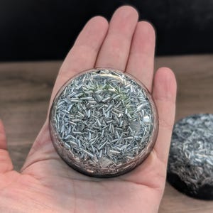 May include: A clear, round orb filled with small, silver-coloured metal pieces. The orb is held in a hand, with another similar object visible in the background. The metal pieces are arranged in a random pattern.