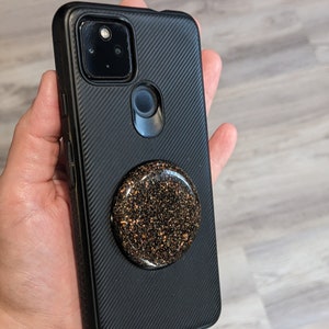 May include: A black phone case with a textured surface and a gold glitter phone grip.