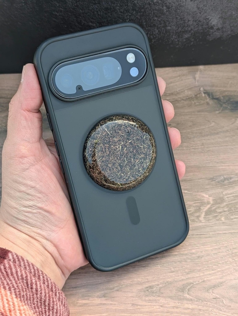 May include: A dark gray phone case with a circular, textured, brown and gold pop socket. The case has a camera cutout and a sleek, minimalist design. The phone case is held in a hand.