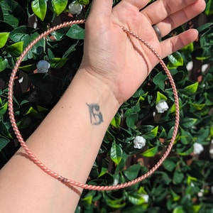 Puede incluir: Un aro de metal de color oro rosa con un diseño de cuerda trenzada. El aro se sostiene en la mano de una persona.