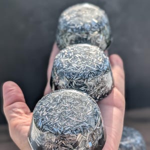 May include: Four clear, dome-shaped objects filled with silver metal shards are held in a hand. The objects have a dark, glossy base and a clear top, creating a layered effect. The background is a blurred, dark grey.