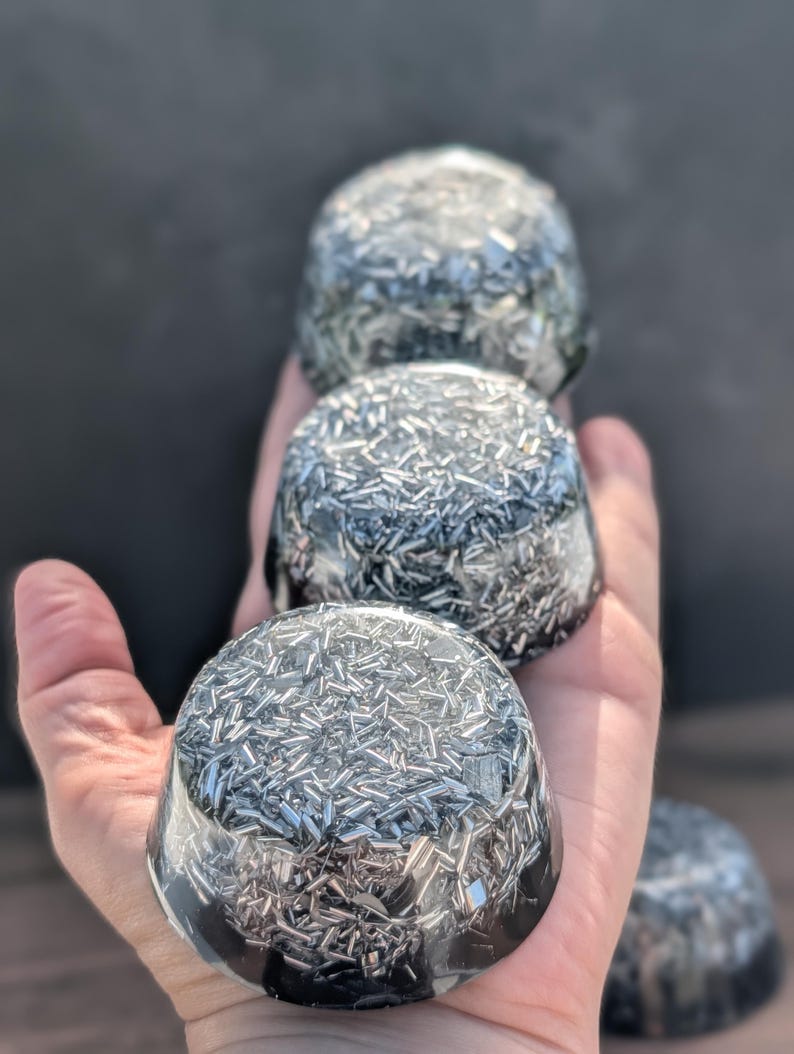 May include: Four clear, dome-shaped objects filled with silver metal shards are held in a hand. The objects have a dark, glossy base and a clear top, creating a layered effect. The background is a blurred, dark grey.