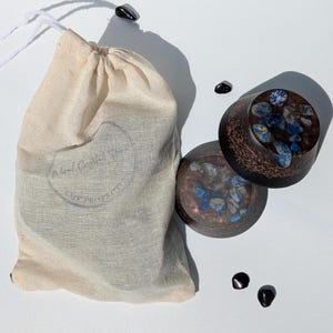 May include: Two dark brown, circular objects with blue and white stones are displayed next to a beige drawstring bag. The objects have a copper-coloured trim. Several small, black, polished stones are scattered around the objects and bag.