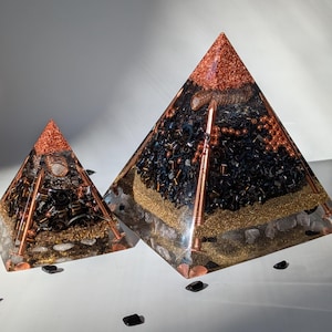 May include: Two black obsidian crystal pyramids with copper wire and beads embedded inside. The larger pyramid is on the right and the smaller pyramid is on the left. Both pyramids have a layer of clear quartz crystals at the base.