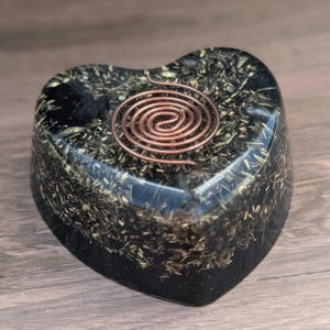 May include: A heart-shaped orgone energy generator. The black resin base is embedded with small, golden-colored metallic particles. A copper wire spiral is centered on the top. The item is displayed on a wooden surface.