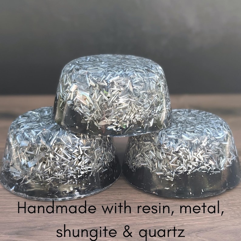 May include: Three dome-shaped objects made with resin, metal, shungite, and quartz. The objects are a dark grey colour with a metallic, reflective appearance. The text "Handmade with resin, metal, shungite & quartz" is at the bottom.