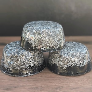 May include: Three black, dome-shaped objects with a silver, glitter-like material embedded throughout. The objects are stacked on top of each other, with the bottom two resting on a wooden surface. The background is a dark grey.